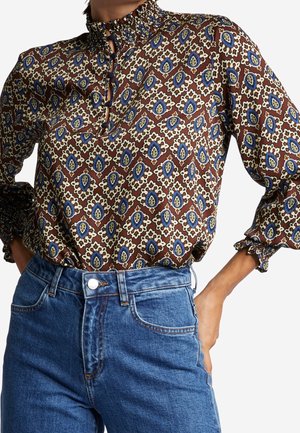 Mujer con blusa marrón estampada con cuello y puños con volantes, combinada con vaqueros azules de cintura alta y una mano en la cadera.