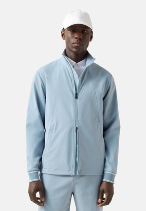 Mand iført lyseblå jakke med lynlås og matchende bukser med hvid kasket, står mod en ensfarvet baggrund.