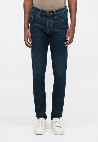 Sötétkék farmernadrág, amely slim fit stílusú, öt zsebbel és arany gombos részletekkel rendelkezik, fehér sportcipővel párosítva. Sima textúra.