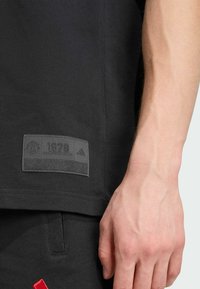 Gros plan sur le bras d'une personne à côté d'un tissu noir avec une étiquette Manchester United 1878 et un détail rouge sur un pantalon noir.