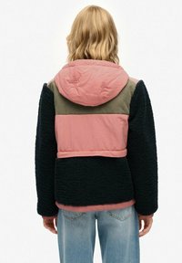 Persoon met schouderlang blond haar, gekleed in een color-block jas met roze, olijfgroene en zwarte secties, en lichtblauwe jeans, met de rug naar de camera gekeerd.
