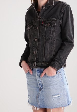 Personne portant une veste en jean noire avec les mains dans les poches et une jupe en jean bleu clair usée avec un ourlet effiloché.