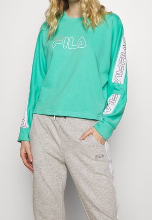 Vrouw draagt een turquoise FILA-trui met logo op de voorkant en mouwen, en grijze FILA-sweatpants met witte strepen aan de zijkant.