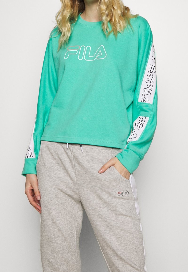 Femme portant un sweat-shirt turquoise FILA avec le logo sur le devant et les manches, et un pantalon de survêtement gris FILA avec des bandes blanches sur les côtés.