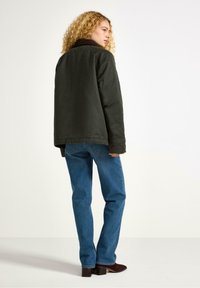Una donna con i capelli ricci si trova di spalle rispetto alla telecamera, indossando una giacca scura e jeans blu, su uno sfondo neutro.