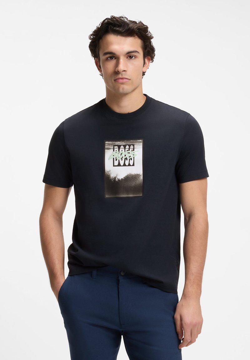 Jeune homme aux cheveux bouclés foncés portant un t-shirt noir à manches courtes avec un graphisme rectangulaire "BOSS" et un pantalon bleu, debout avec une main dans la poche.
