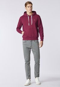 Felpa bordeaux con tasca frontale e cordini, abbinata a jeans slim-fit grigi e sneakers bianche su uno sfondo neutro.