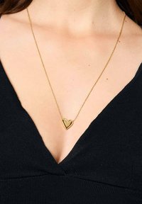 Złoty naszyjnik z zawieszką w kształcie serca na cienkim łańcuszku, z dwukolorowym wzorem składającym się z wypełnionego i konturowego serca. Nosi się go na czarnej odzieży.