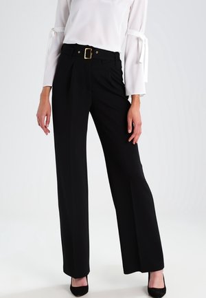 Kobieta ubrana w czarne spodnie z szerokimi nogawkami i wysokim stanem, ozdobione złotą klamrą paska, w połączeniu z białą, długorękawową bluzką z wiązanymi rękawami oraz czarnymi szpilkami.