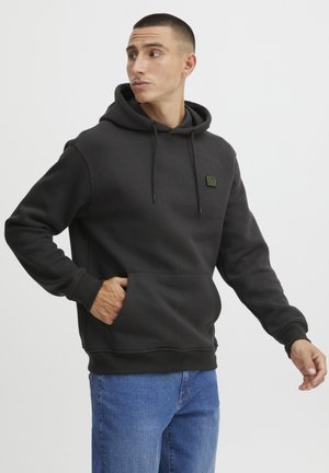 Black hoodie made of soft fabric, featuring a kangaroo pocket, drawstring hood, and a green logo patch on the chest. Worn with blue jeans.