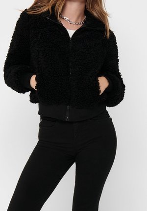 Frau trägt eine schwarze, strukturierte Jacke mit Reißverschluss und den Händen in den Taschen, kombiniert mit schwarzer Hose und einer silbernen Ketten-Halskette.