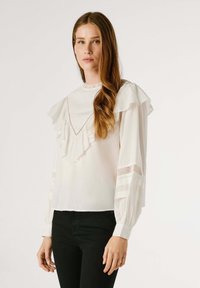 Blusa bianca con scollo arricciato, dettagli in pizzo e maniche lunghe. Presenta accenni in rete trasparente e una vestibilità dritta. Indossata con pantaloni neri.
