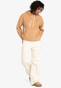 Tan fleece hoodie with front pocket and white drawstrings, paired with off-white wide-leg pants and brown shoes with textured soles.