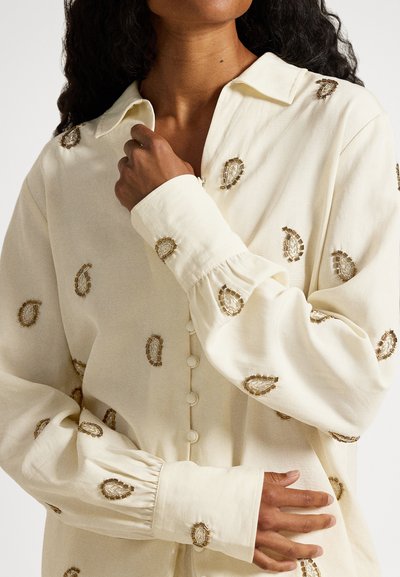 Cremefarbene Hemdbluse mit dekorativer paisley-goldfarbener Stickerei und gesammelten Ärmelsäumen. Weicher, strukturierter Stoff.