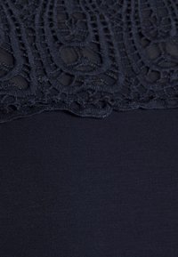 Zblízka tmavá tkanina s propracovanou krajkovou výšivkou v horní části a hladkým textilem v dolní části.