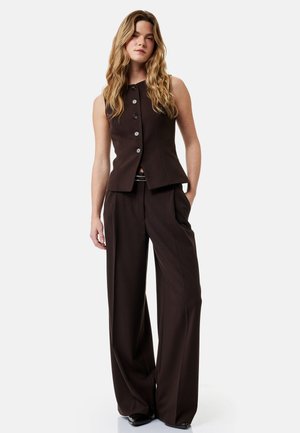 Vrouw met lang golvend haar draagt een mouwloos zwart gilet met knopen en hoge taille, wijde zwarte broek, staand met één hand in de zak.