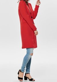 Blazer largo rojo de textura suave, con cierre de un solo botón y dos bolsillos laterales, llevado sobre jeans rasgados de color azul claro y sandalias negras de tacón alto.