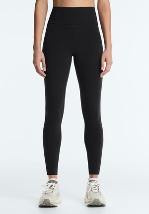 Nő fekete, magas derekú leggingset és fehér tornacipőt visel, egyszerű világos háttér előtt áll.