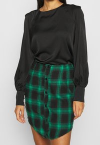 Musta satiini pitkähiha blouse pyöreällä kaula-aukolla, yhdistettynä vihreään ja mustaan ruudulliseen hameeseen, jossa on etupainikkeet ja risat helma.