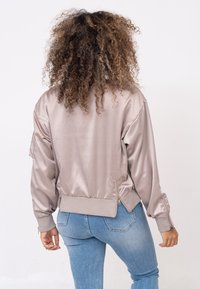 Satijnen bomberjack in licht taupe, met ribgebreide boorden en zoom, ritssluiting zijzakken en een gladde textuur, gedragen over blauwe jeans.