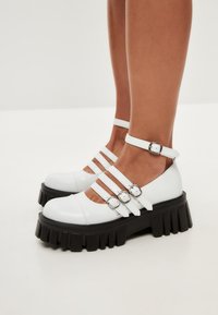 Scarpe platform in pelle bianca con tre dettagli a cinturino e fibbia regolabile alla caviglia, dotate di una suola in gomma nera testurizzata.