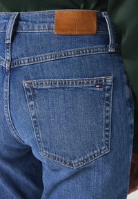 Plave traperice s kožnom etiketom na struku oznake "TOMMY HILFIGER". Imaju klasičan kroj s stražnjim džepovima i vidljivim šavovima.