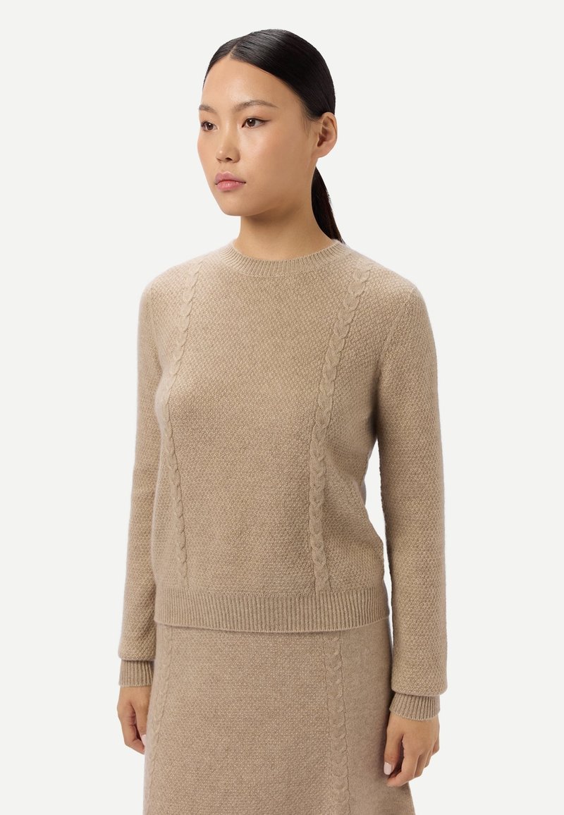 Frau trägt beige gestrickten Pullover und passenden Rock mit Zopfmuster-Details, steht vor weißem Hintergrund und blickt zur Seite.