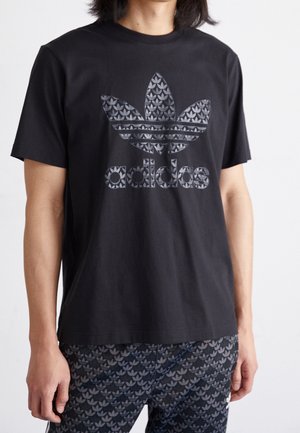 Personne portant un t-shirt Adidas noir avec un grand logo trèfle à motifs et un short assorti, debout sur un fond blanc.