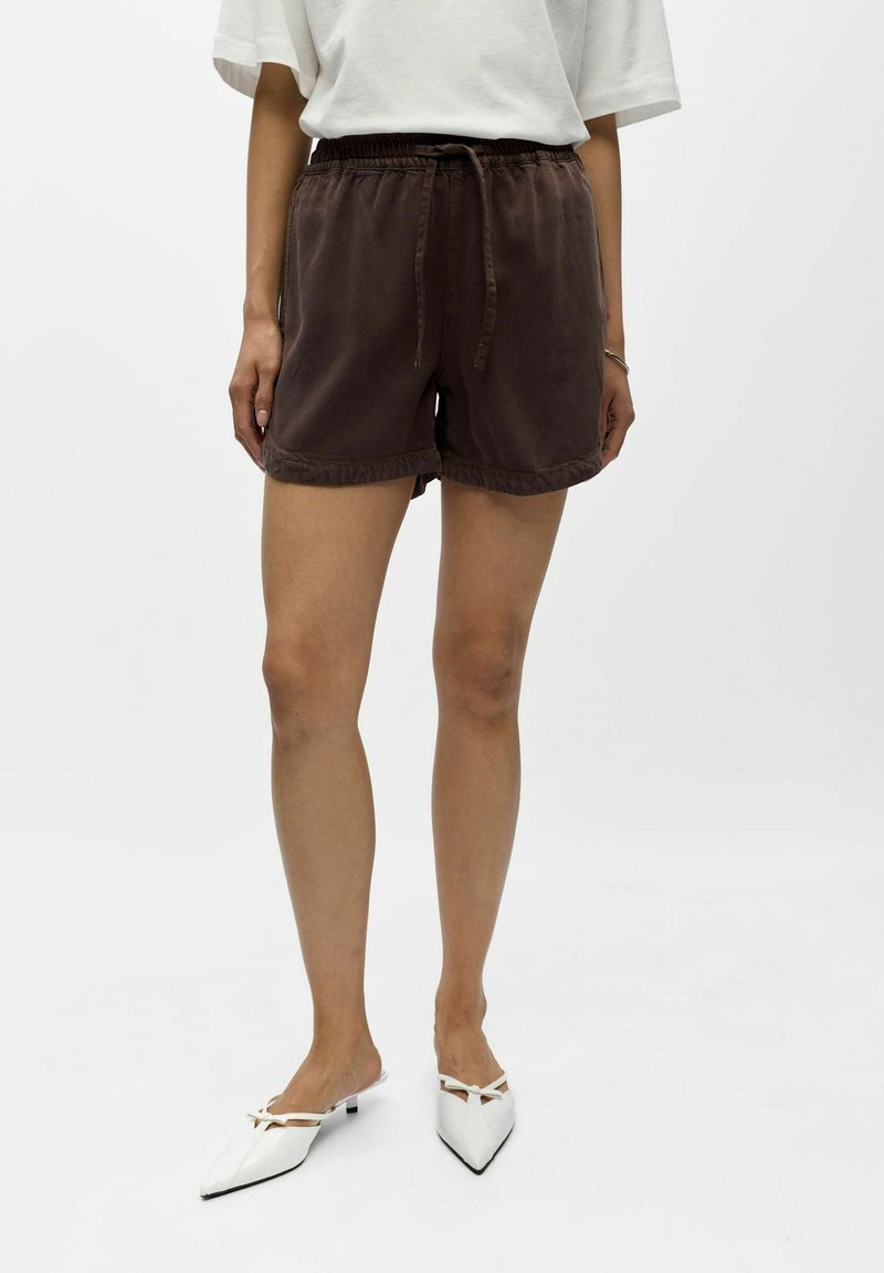 Personne portant un short marron à cordon, une chemise blanche à manches courtes et des mules blanches à talons pointus sur un fond uni.