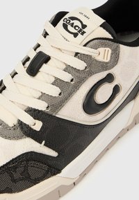 Texturált törtfehér felsőrészű sneaker, szürke részletekkel, fekete bőrdíszítéssel, bézs talppal, fekete logóval és krémszínű fűzőkkel.