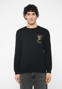 Νεαρός άνδρας με σγουρά μαλλιά που φοράει μαύρο πουλόβερ με χρυσό γραφικό άλογο και κείμενο στο στήθος, συνδυασμένο με σκούρες γκρι τζην, κοιτάζοντας μπροστά.