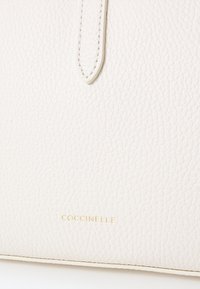 Torba Coccinelle od teksturirane ecru kože s glatkom trakom. Ima zlatni otisnuti logo na dnu. Minimalistički dizajn.