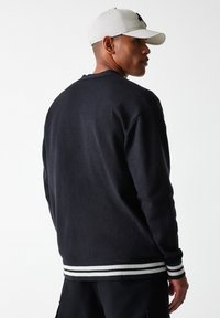 Pull noir avec un col rond, des poignets et un ourlet côtelés arborant des rayures blanches, fabriqué en tissu doux, vu de dos avec une casquette beige.