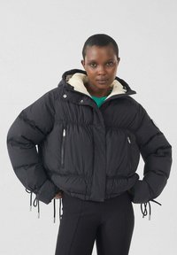 Zwarte donsjas met een crèmekleurige capuchon, voorzien van twee voorzakken met rits, elastische manchetten en verstelbare trekkoorden in de taille.