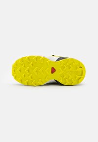 Running shoe featuring a yellow outsole with deep tread patterns, white upper, black accents, and a red logo detail.