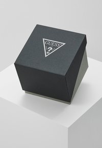 Caja de regalo negra, texturada, con una tapa triangular; presenta un logo plateado en la parte superior. La base es de un gris más claro. Posicionada sobre un pedestal blanco.