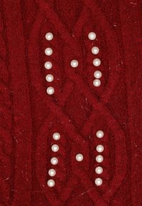 Piros kötött anyag emelt kábelmintázattal, díszítő fehér gyöngyökkel, amelyek az "M" és "H" betűk formájában vannak elrendezve.