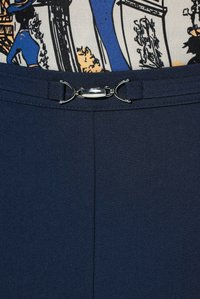 Marineblauwe broek met een zilverkleurige metalen gespriem, gedragen met een patroonhemd met blauwe en beige abstracte ontwerpen.