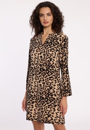 Vrouw in een langwerpige, knielange jurk met een beige en zwarte luipaardprint, staande tegen een effen achtergrond.