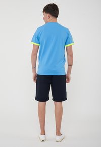 T-shirt di cotone azzurro chiaro con dettagli giallo neon alle maniche, abbinato a pantaloni corti blu navy e scarpe bianche. Aspetto pulito e informale visto da dietro.
