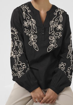 Czarna bluzka z długim rękawem z kremowym haftem kwiatowym na klatce piersiowej i rękawach, noszona przez siedzącą osobę z rękami spoczywającymi na beżowych spodniach.