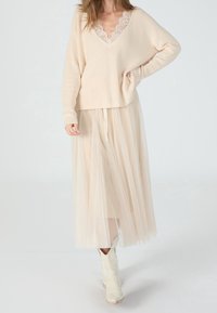 Pull en maille beige avec un col en V orné de dentelle, associé à une longue jupe en tulle beige transparent et des bottines blanches.