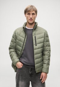 Φούτερ μπουφάν σε ανοιχτό ελαιόχρωμο με μπροστινό φερμουάρ, ψηλό γιακά και υφή με τσάκιση. Συνδυασμένο με σκούρο γκρι πουλόβερ και τζιν.