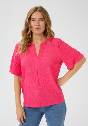 Blouse rose texturée à manches courtes avec un décolleté en V. Présente une coupe décontractée et des détails de fronces subtils. Associée à un jean en denim bleu.