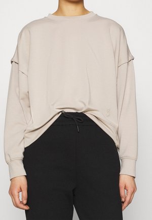 Personne portant une chemise beige ample à manches longues rentrée dans un pantalon noir à cordon, vue des épaules aux hanches.