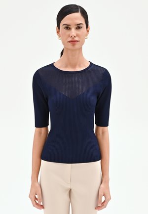 Donna con capelli scuri raccolti in una coda bassa, indossa una maglia blu navy a maniche corte con motivo a coste e inserto trasparente sullo scollo, abbinata a pantaloni beige, in piedi davanti a uno sfondo bianco.