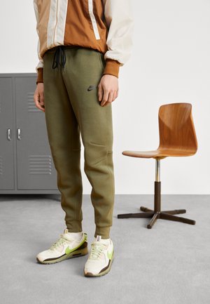 Person trägt eine braune und cremefarbene Jacke, olivgrüne Nike-Sweatpants und weiße Turnschuhe mit grünen Akzenten, steht neben einem drehbaren Holzstuhl.