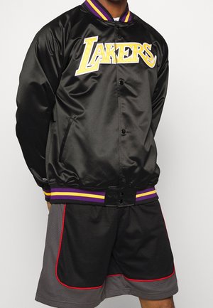 Person trägt schwarze Satin-Lakers-Jacke mit lila und gelbem Besatz sowie zweifarbige schwarz-graue Basketballshorts mit roter Paspelierung.