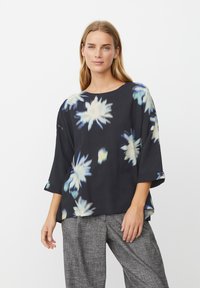 Sort blomsterbluse med blå og hvide abstrakte blomstermønstre, vide ærmer, rund halsudskæring og let stof, kombineret med grå teksturerede bukser.