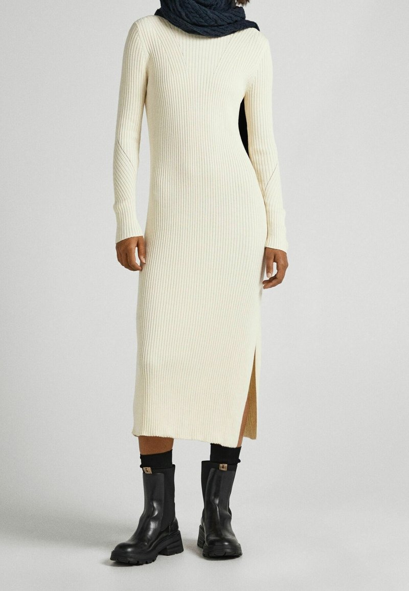 Stickad krämfärgad klänning med ribbad textur, långa ärmar, sidoklyv, tillsammans med svarta ankelskängor och en mörk scarf runt halsen.